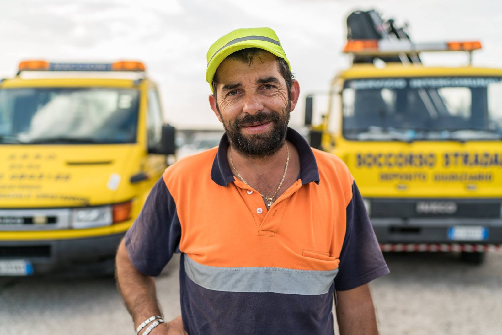 Operatore in polo arancio-blu e cappellino fluo davanti ai carroattrezzi gialli A.S.S.O. con scritta Soccorso Stradale