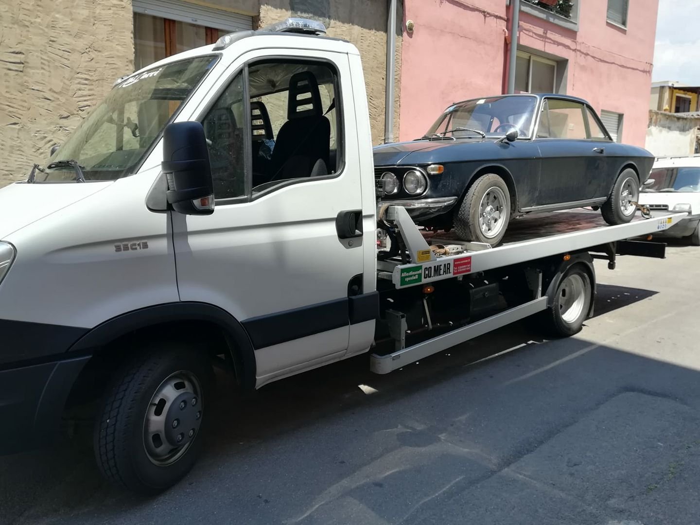 Carroattrezzi bianco A.S.S.O. con pianale CO.ME.AR. che trasporta una coupé d'epoca blu scuro in una via cittadina